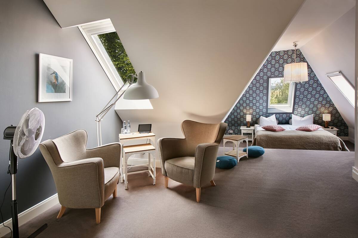 A bedroom with a sloped ceiling, a desk, two chairs, a bed, and a standing fan.