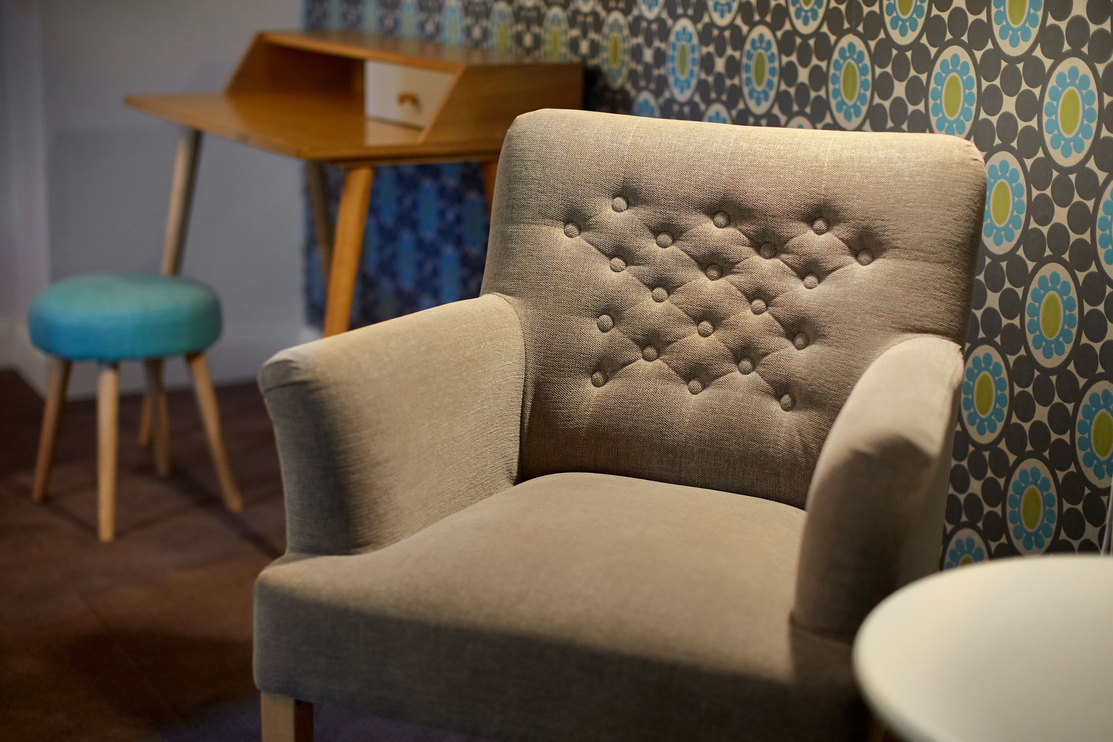 A beige armchair with a tufted backrest, a wooden side table, and a blue cushioned stool in a room with patterned wallpaper.