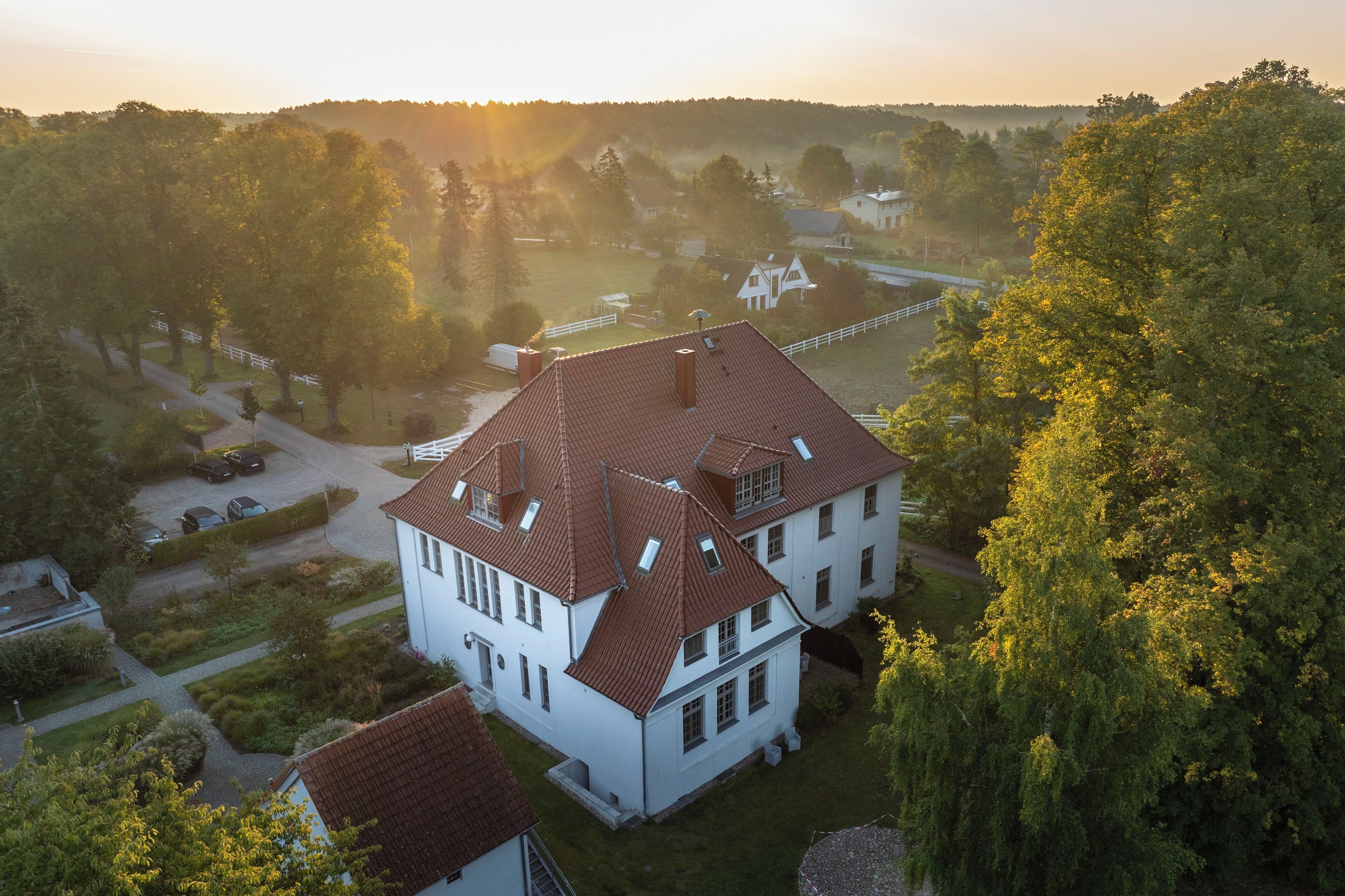 A large white building with a red roof, surrounded by greenery and a clear sky.