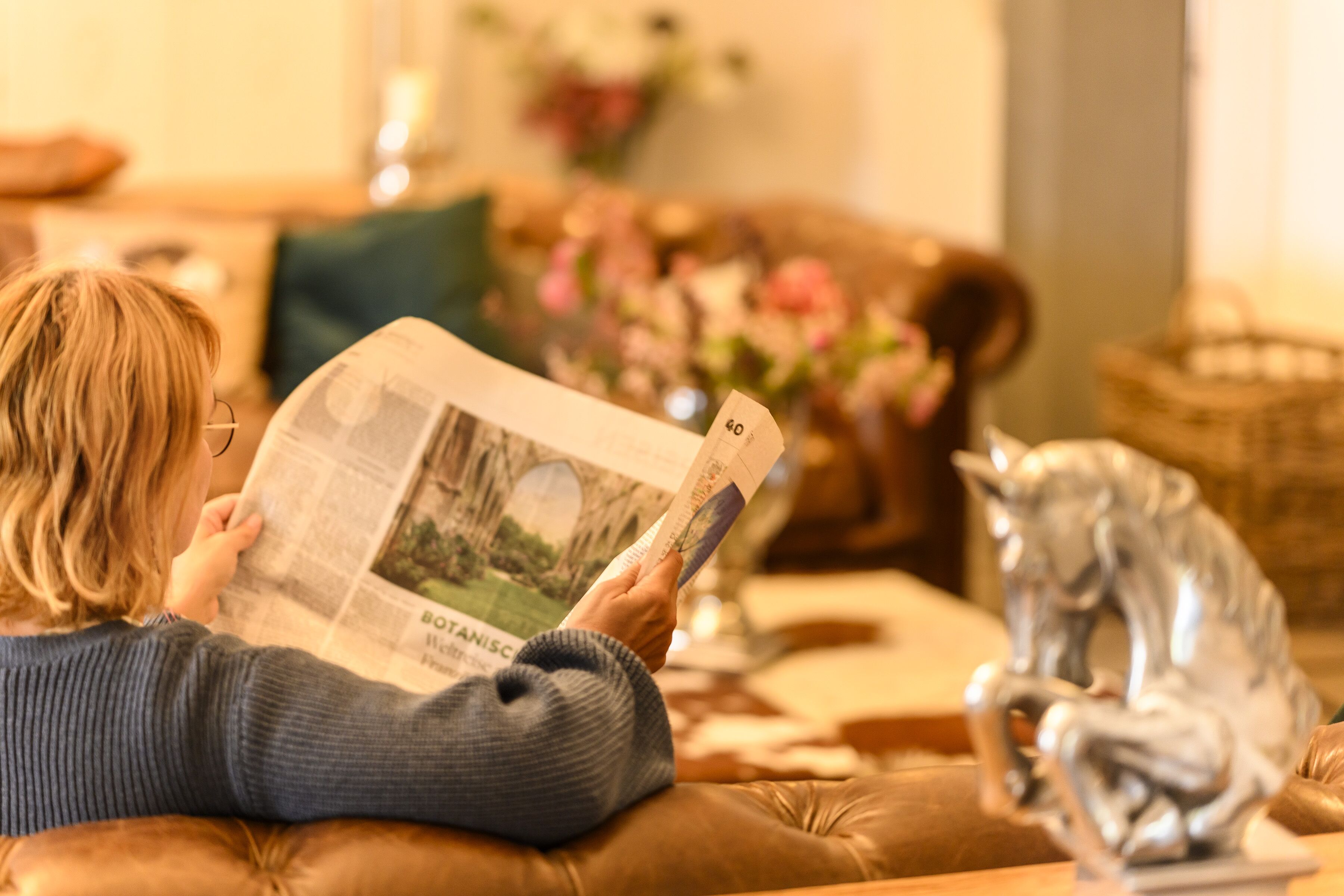 Una persona leyendo un periódico en una sala de estar acogedora con un sofá de cuero, una estatua de plata y un jarrón con flores.