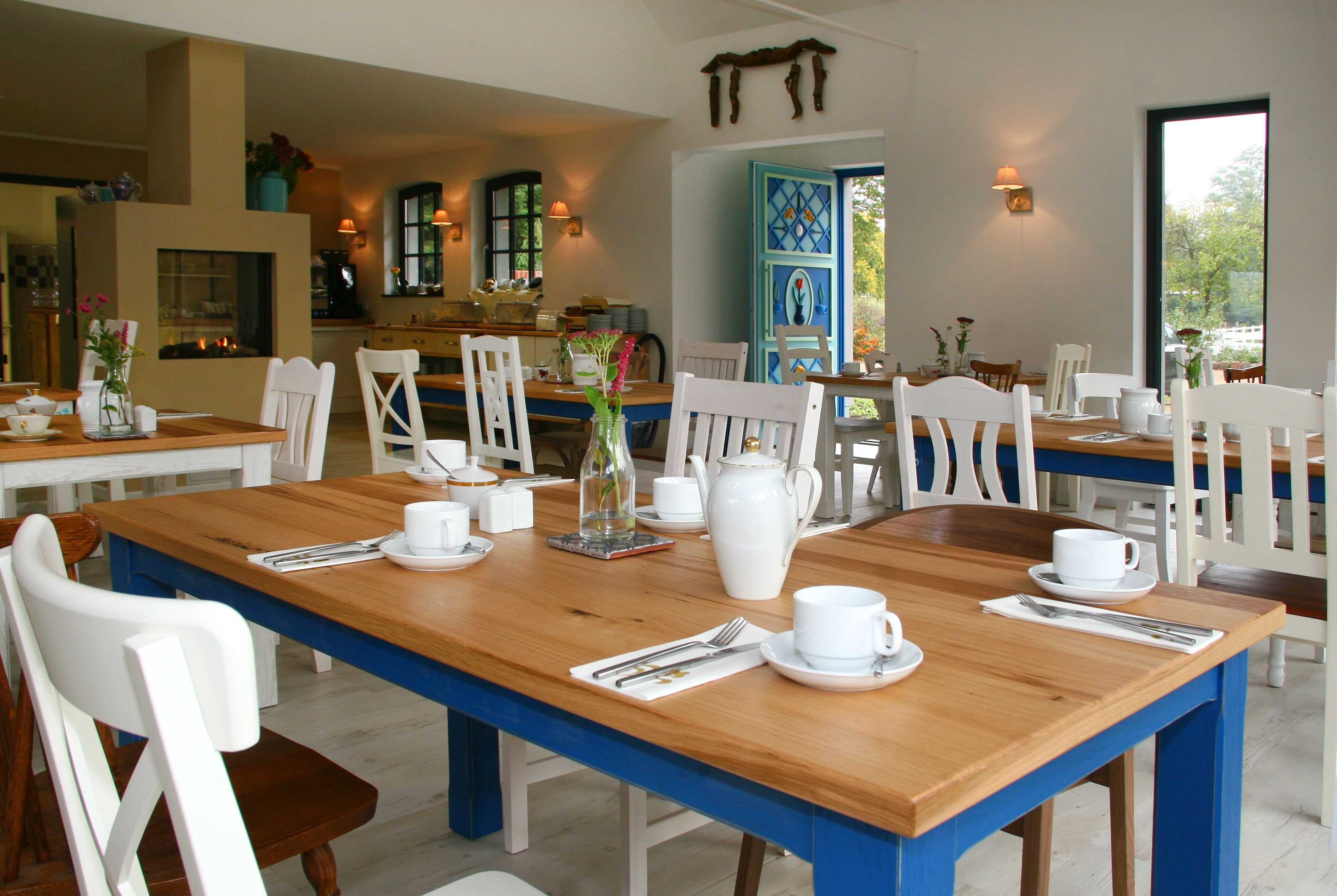 Un coin repas avec des tables en bois et des chaises blanches, un vase rempli de fleurs et, au fond, une porte bleue.