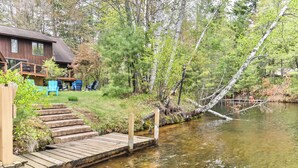 A wooden dock extending into a calm river, surrounded by lush greenery and a wooden cabin with outdoor seating. - Gorgeous large lakefront home with firepit and a private dock! (ST GERMAIN)