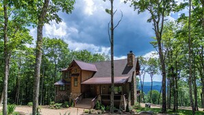 A rustic cabin with a stone chimney, wooden exterior, and a porch with steps leading to a deck.