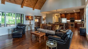 A spacious living room with a high ceiling, wooden beams, a large sofa, a coffee table, and a view of the outdoors through large windows.