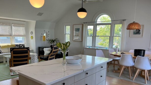 A modern kitchen with a marble countertop, wooden cabinets, and a central island. There are two pendant lights, a dining area with a table and chairs, and a large window providing natural light. - Open Floor Plan, Family Friendly, Waterview Home  (Narragansett)