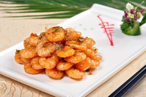 A white rectangular plate with a pile of cooked shrimp garnished with herbs, placed on a table with a decorative red symbol and a small floral arrangement. - Shanghai Xinjiangnan Hotel (Shanghai)