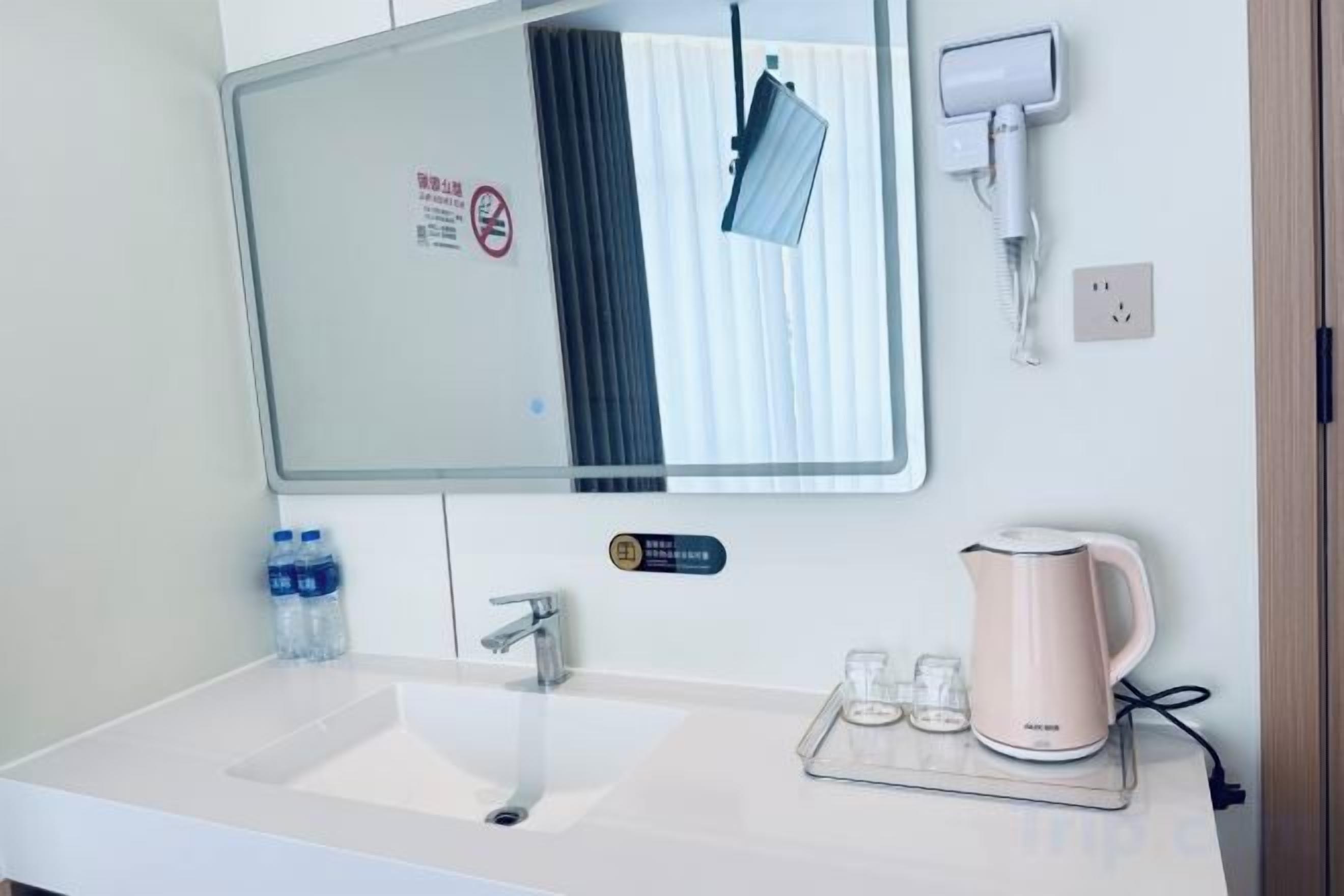 A hotel bathroom with a sink, mirror, towel dispenser, and a kettle on the counter.