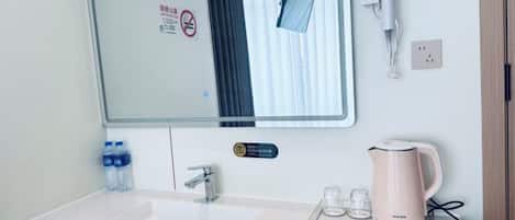 A hotel bathroom with a sink, mirror, towel dispenser, and a kettle on the counter.