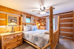 A log cabin bedroom with a bed, bedside lamps, a wooden dresser, a ceiling fan, and a window with curtains.