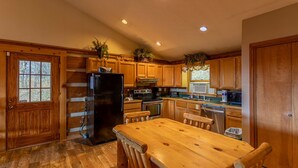 A kitchen with wooden cabinets, a black refrigerator, a wooden dining table with chairs, and a window with a view of trees. - Silverleaf - 30 Mile View, Hot Tub, Close to ASU (Zionville)