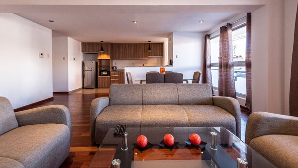 Una sala de estar moderna con un sofá gris, una mesa de centro con manzanas rojas y una cocina al fondo.