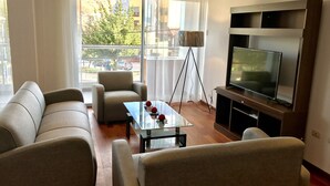 Una sala de estar moderna con un juego de sofá, una mesa de centro de vidrio con adornos, una lámpara de pie y un televisor de pantalla plana sobre un soporte de madera.