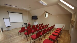 Uma sala de conferências com fileiras de cadeiras vermelhas, uma lousa, uma tela de projetor e uma televisão.