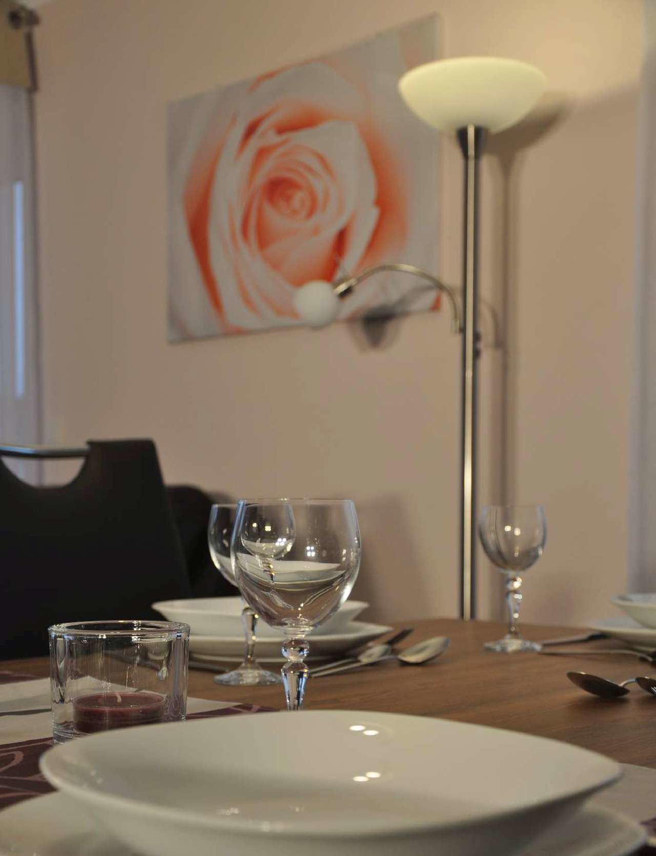 A dining table with a white plate, a wine glass, and a candle. There is a painting of a rose on the wall behind the table.