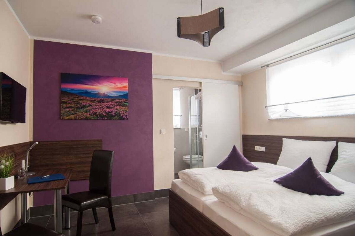 A modern hotel room with a bed, a desk, a chair, a painting, and a bathroom visible through a glass door.