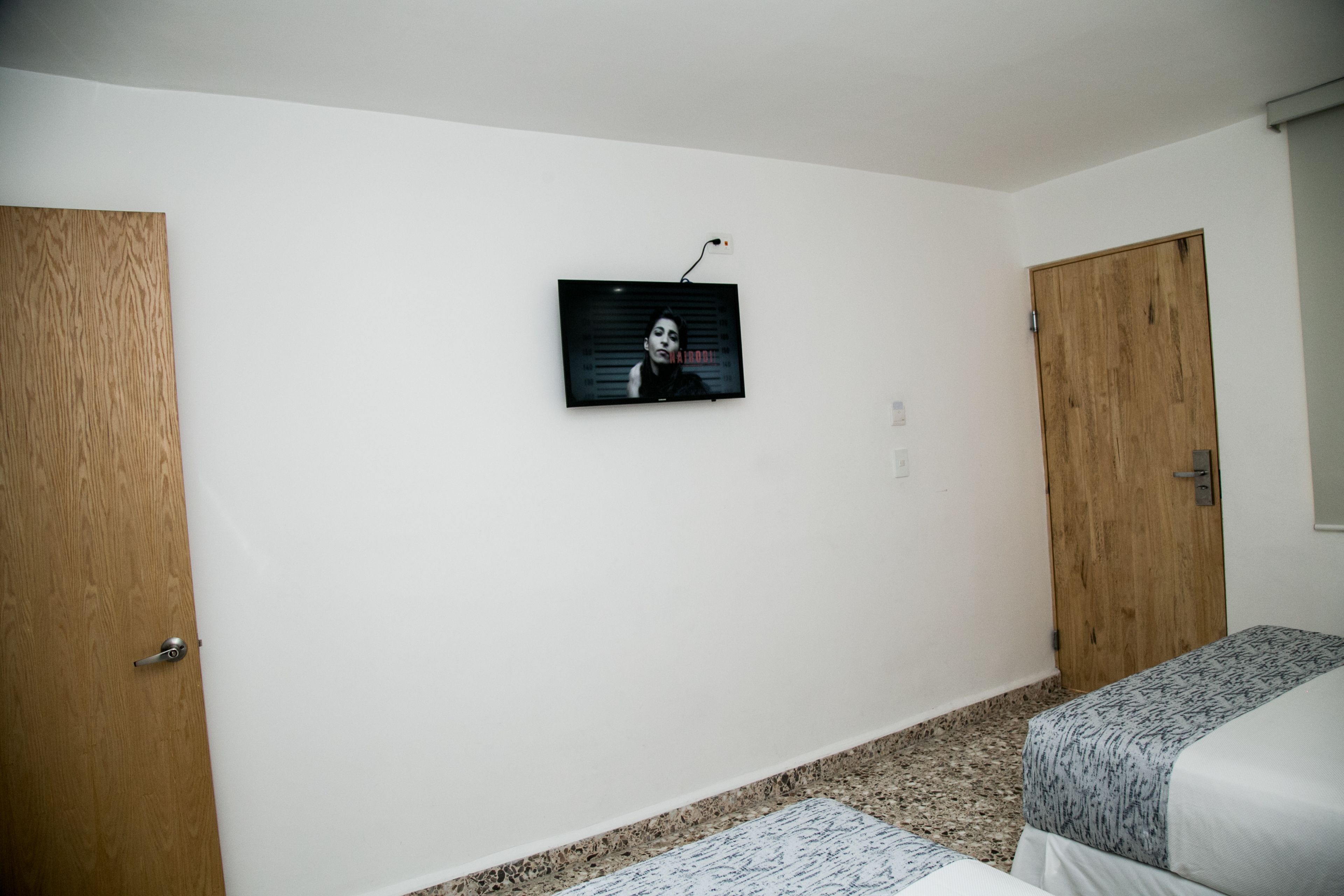 A room with a television mounted on the wall, two wooden doors, and two beds with patterned bedding.