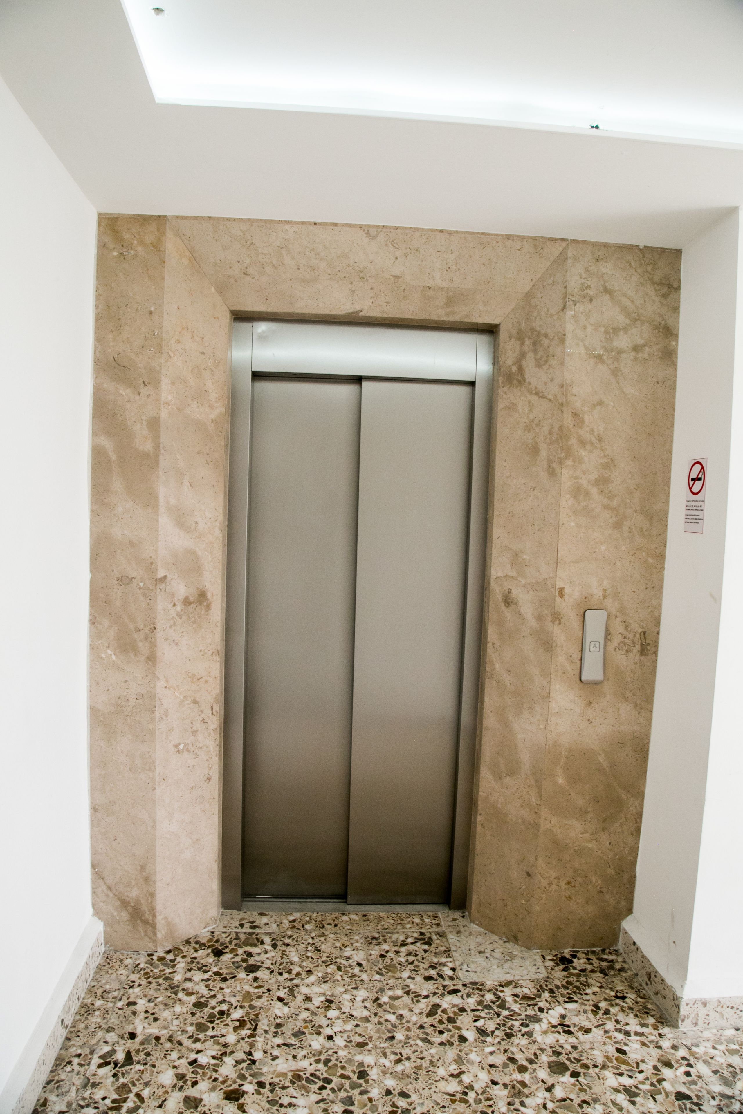 En elevator i en gang med et plettet gulv og lyse beige vægge.