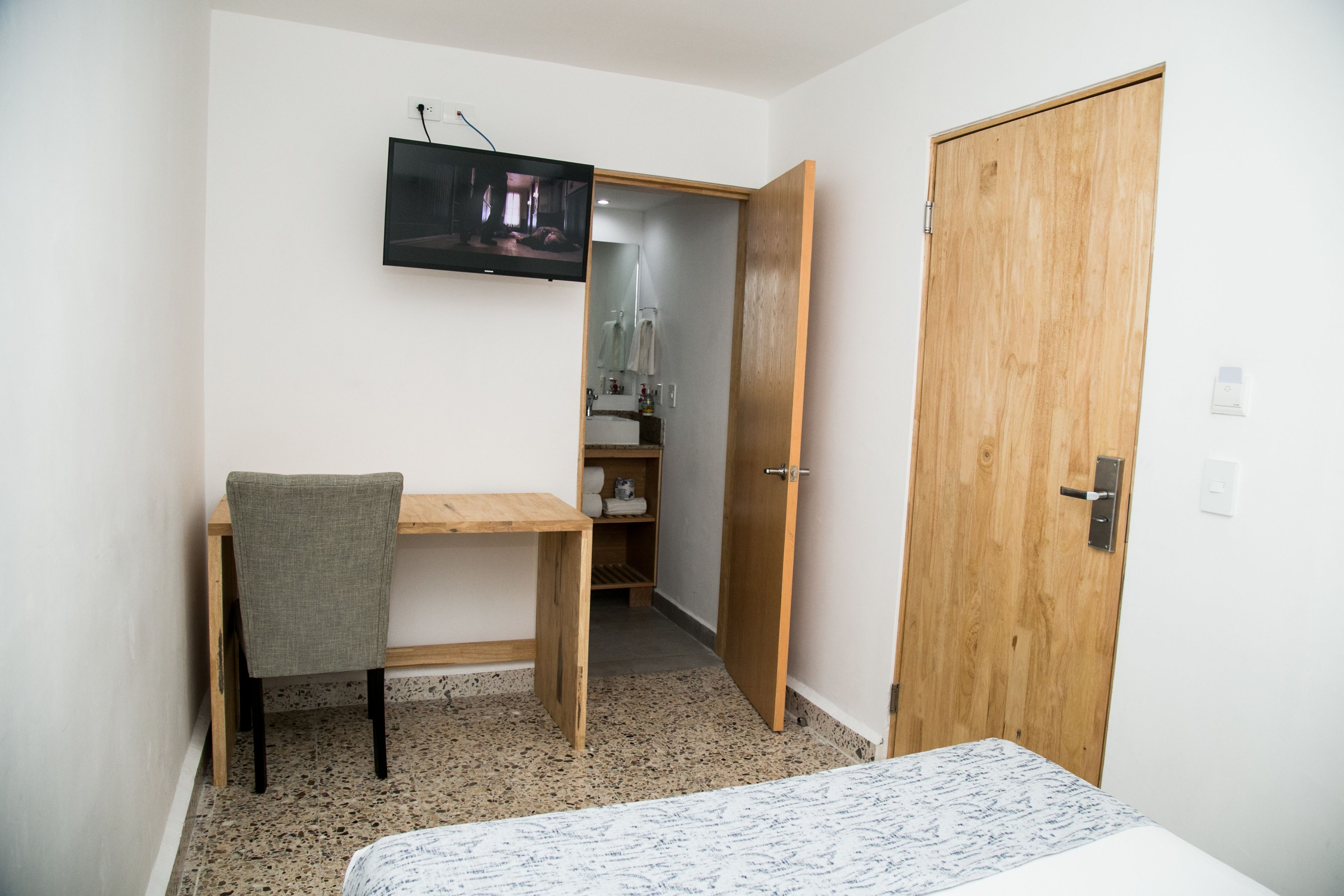 A small room with a wooden desk, a chair, a bed, a television mounted on the wall, and a wooden door.