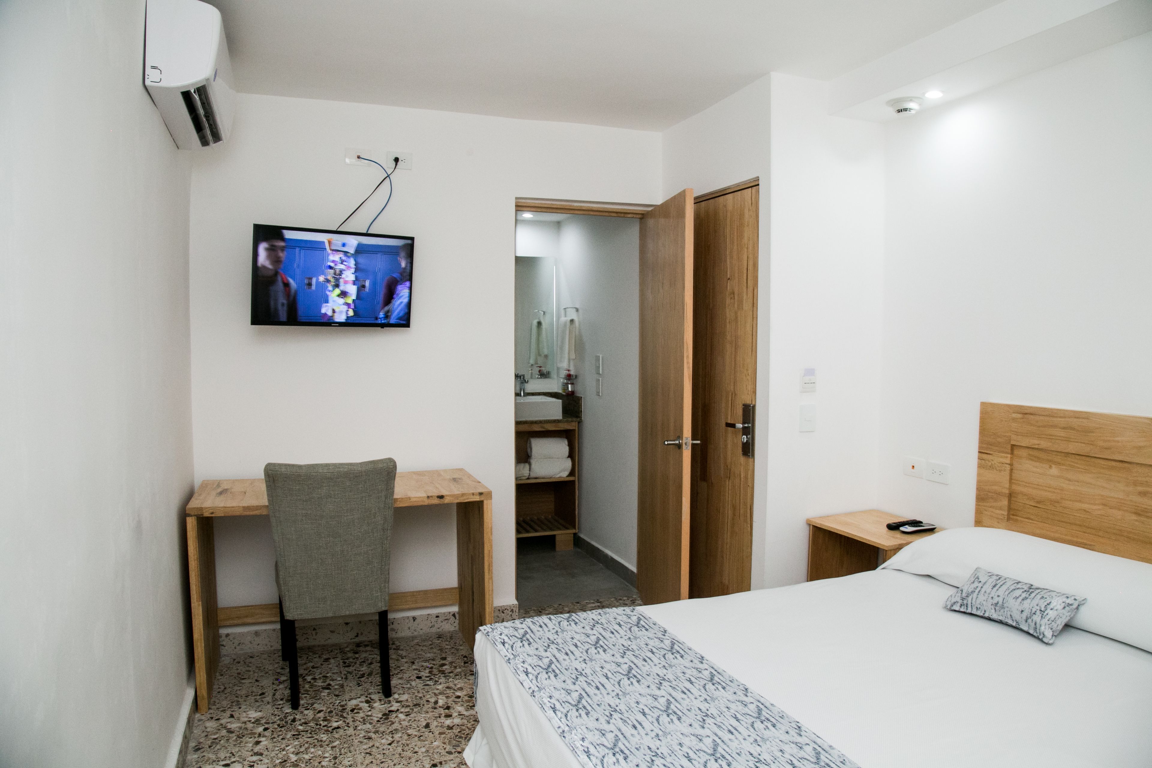 Habitación de hotel con cama, escritorio, silla, televisor, armario y baño visible a través de una puerta abierta.