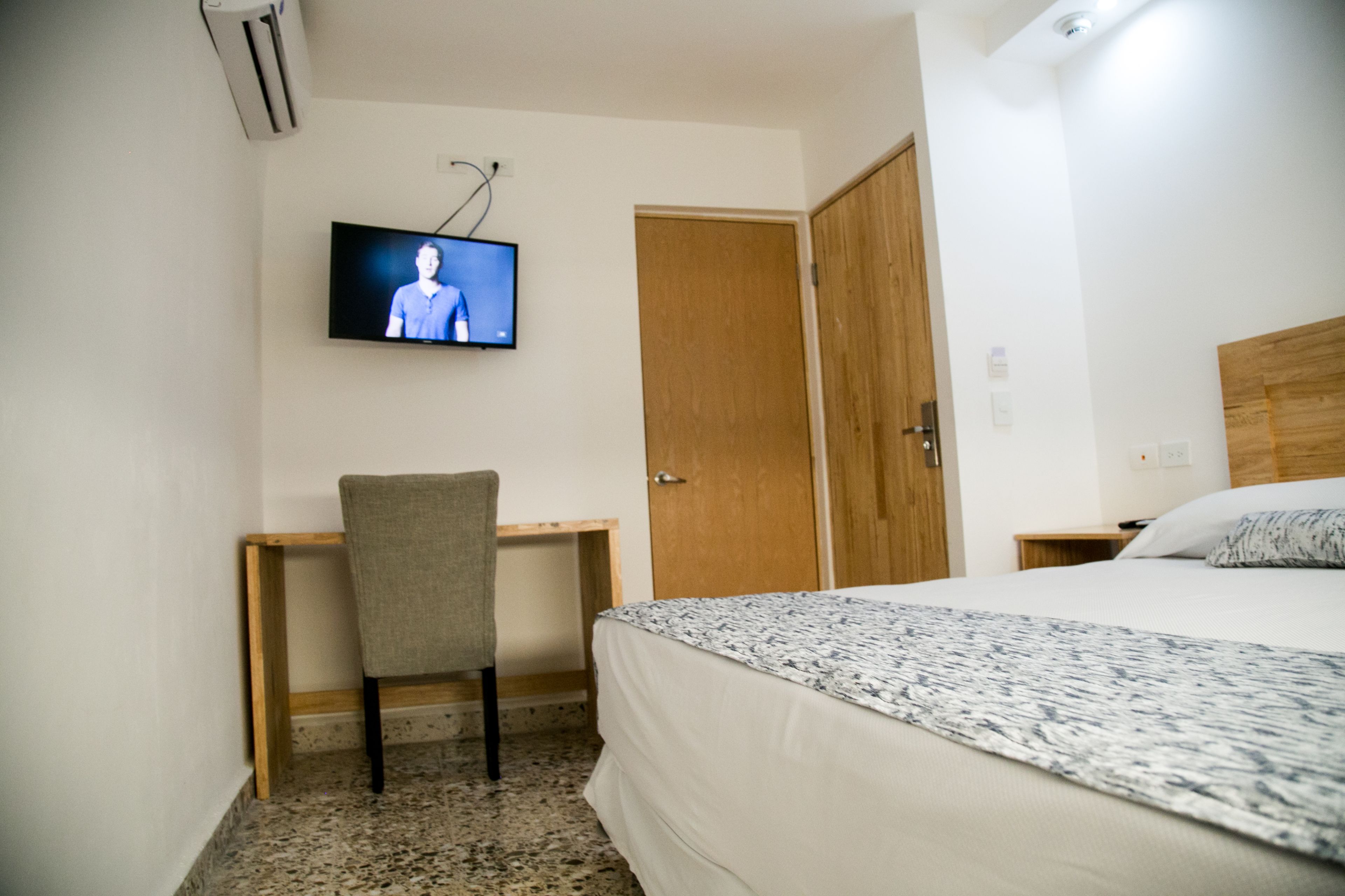 A hotel room with a bed, a desk with a chair, a television mounted on the wall, and a wooden door.