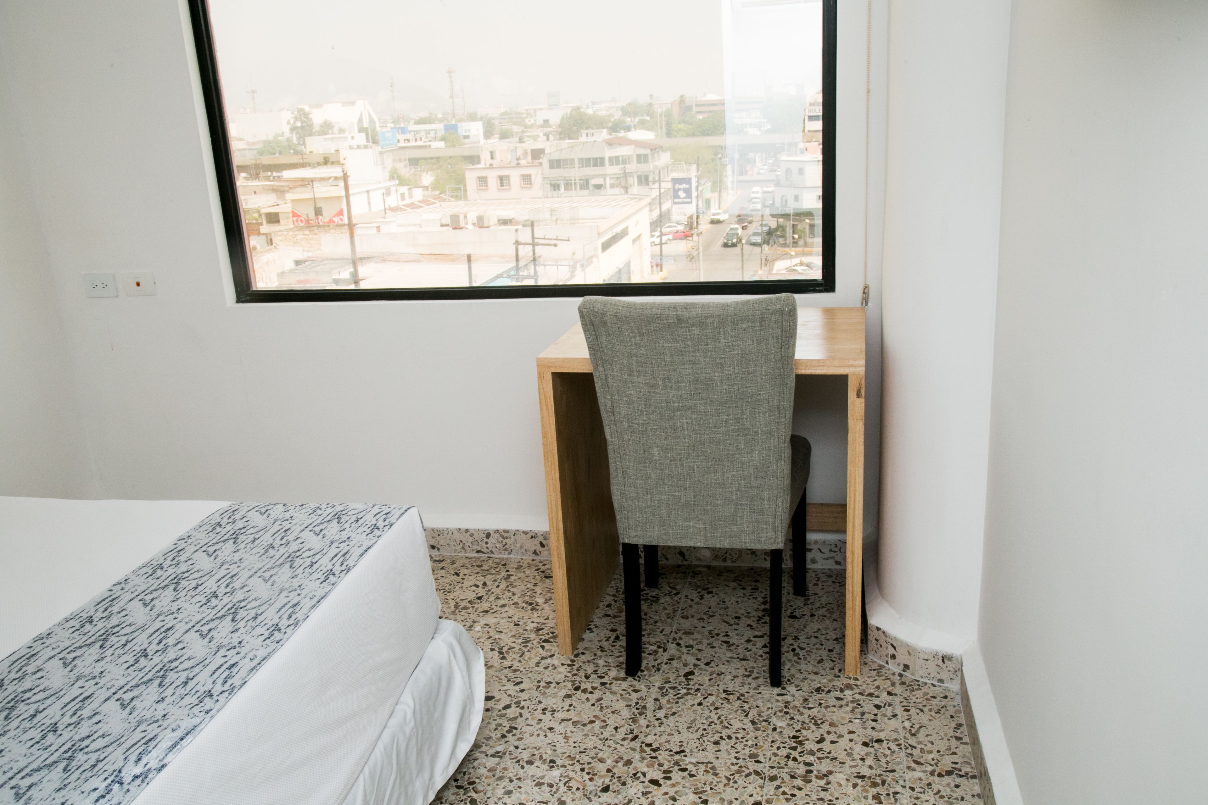 A bedroom with a bed, a desk, and a chair, a window with a city view, and a tiled floor.