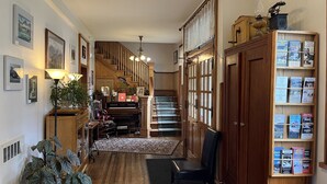 A hallway with a piano, a bookshelf with travel brochures, a staircase, and a potted plant. - At the White House (Skagway)