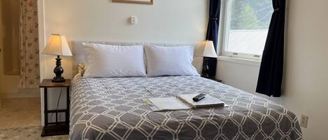 Ein ordentlich bezogenes Bett mit einem geometrisch gemusterten Überwurf, zwei Nachttischlampen, ein gerahmtes Bild an der Wand und ein Fenster mit Vorhängen.