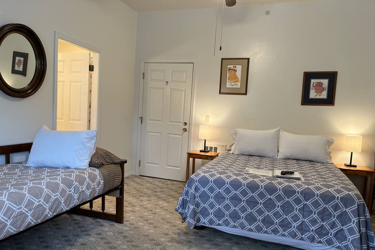 A hotel room with two beds, a door, a wall clock, and framed pictures.