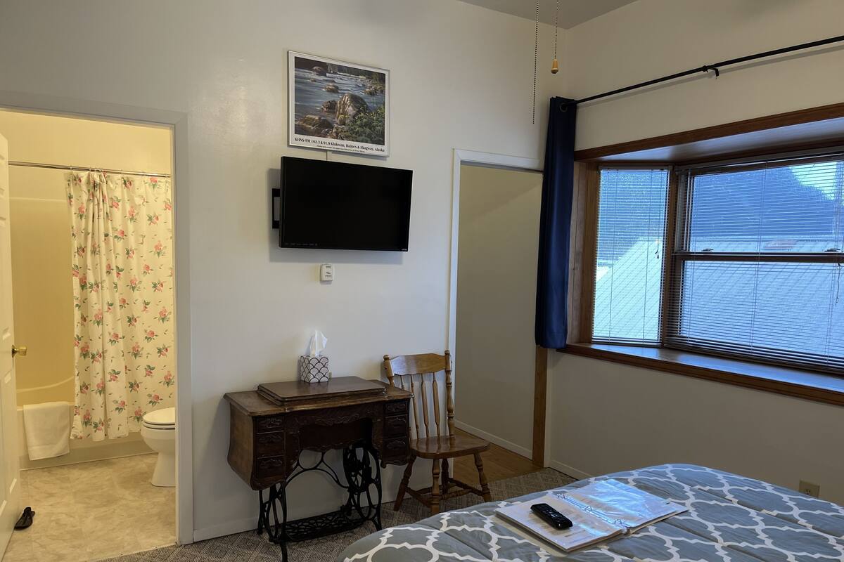 A bedroom with a bed, a desk, a chair, a television, and a window with blinds.
