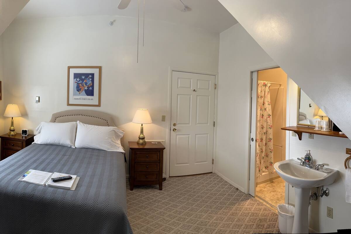 A bedroom with a bed, two bedside lamps, a nightstand, a door, a bathroom with a sink and mirror, and a framed picture on the wall.