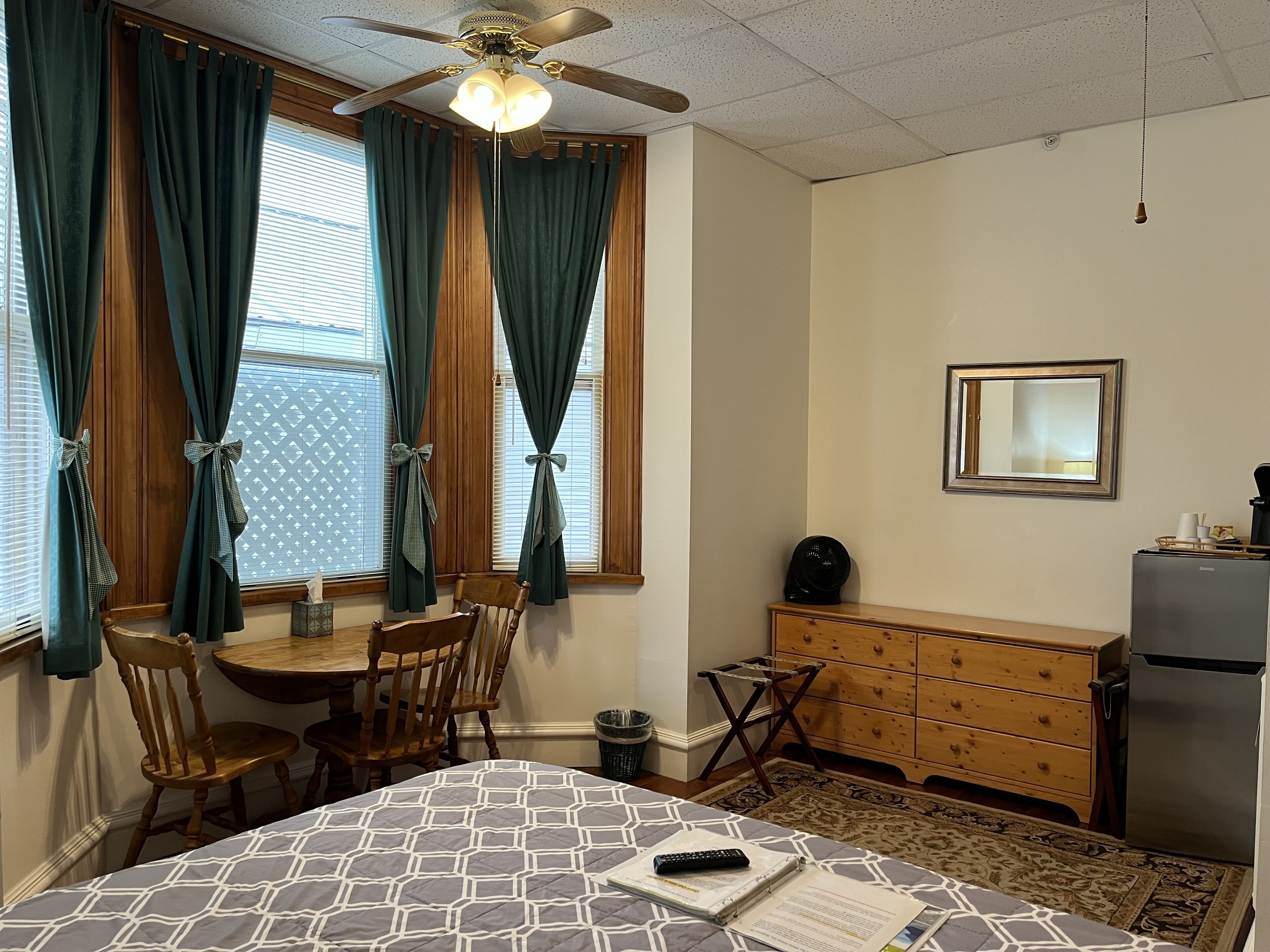 A compact hotel room with a bed, a desk, a chair, a small table, a refrigerator, a ceiling fan, and a window with curtains.
