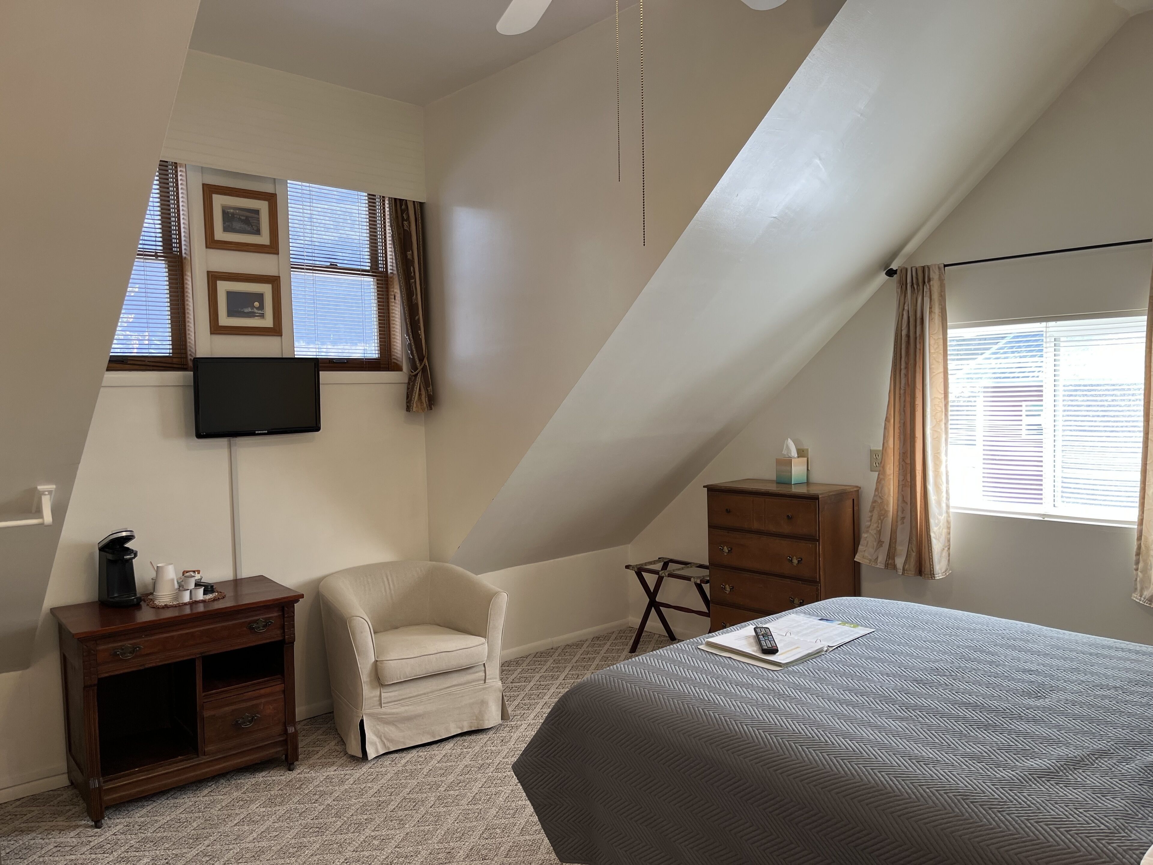A bedroom with a sloped ceiling, a bed with a patterned bedspread, a chair, a small wooden dresser, a television mounted on the wall, and a window with blinds.