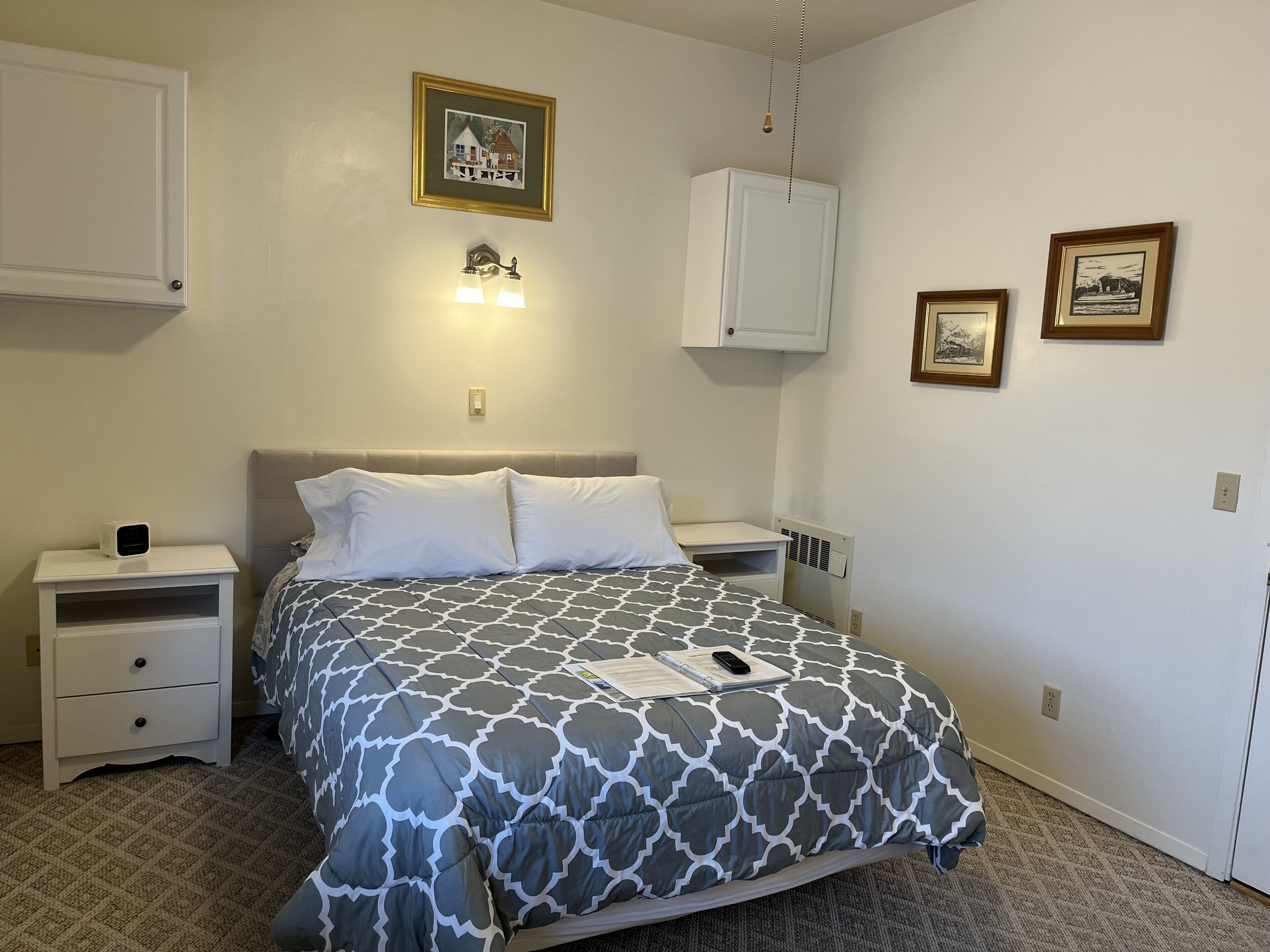 A neatly arranged bedroom with a bed, bedside tables, a nightstand, a wall-mounted light fixture, and framed pictures.