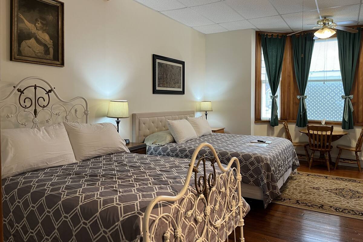 A hotel room with two beds, a wooden ceiling, a ceiling fan, a window with curtains, and a small table with chairs.