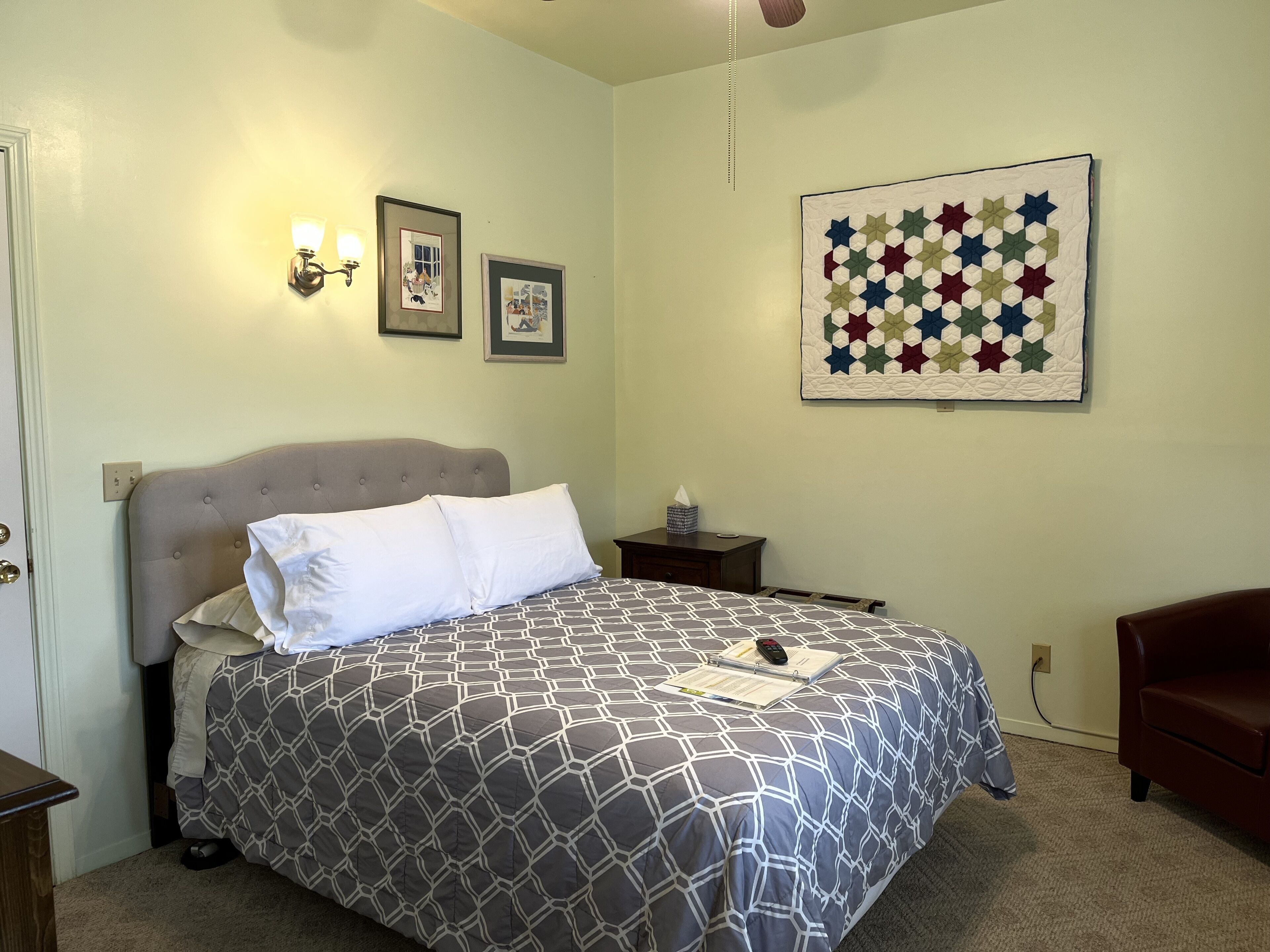 A neatly made bed with a geometric patterned bedspread, a bedside table with a lamp, and a wall-mounted quilt.