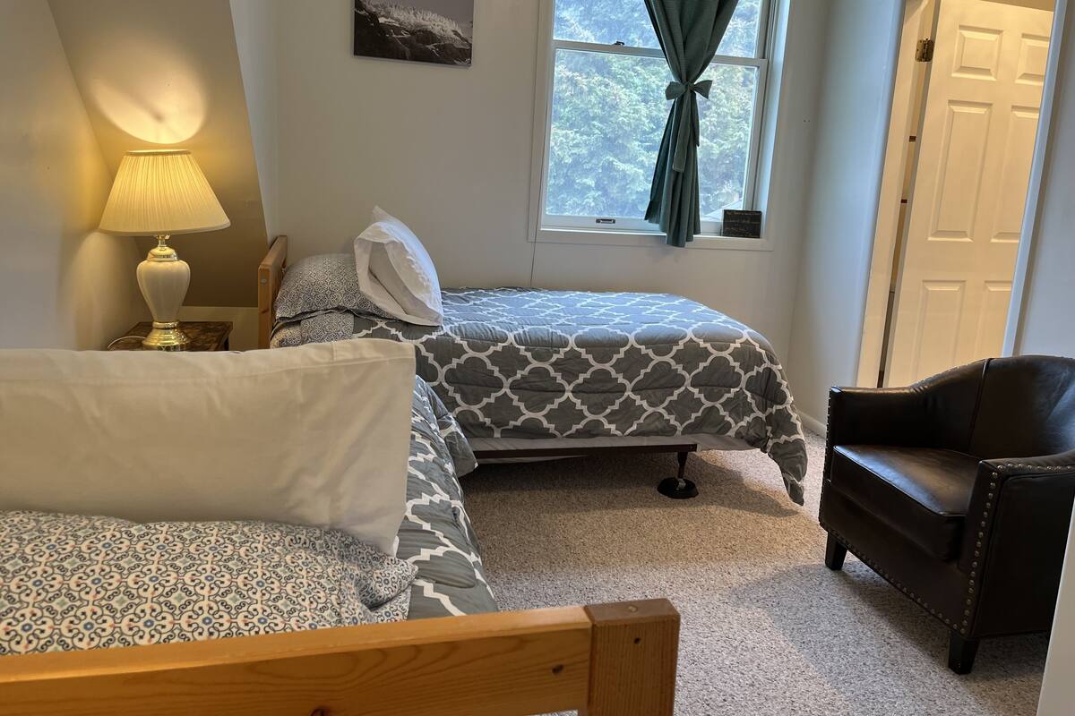 A bedroom with two beds, a lamp, a chair, a window with curtains, and a picture on the wall.