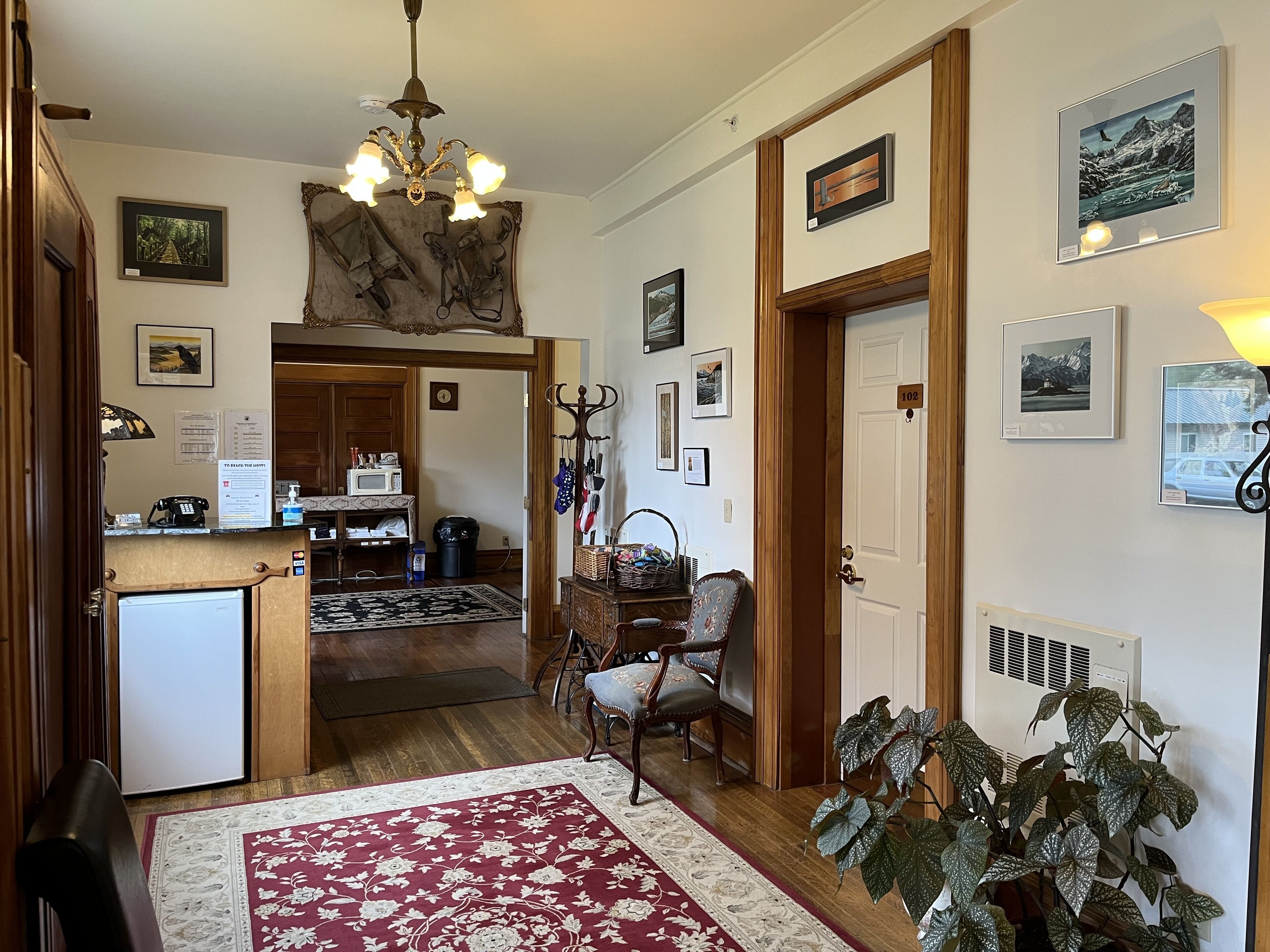 A cozy room with a wooden floor, a red and white patterned rug, a chandelier, and various framed pictures on the walls.