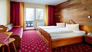 A hotel room with a wooden bed, a desk with a chair, a small table, and a view through a window. - Hotel Wieser (Mittersill)