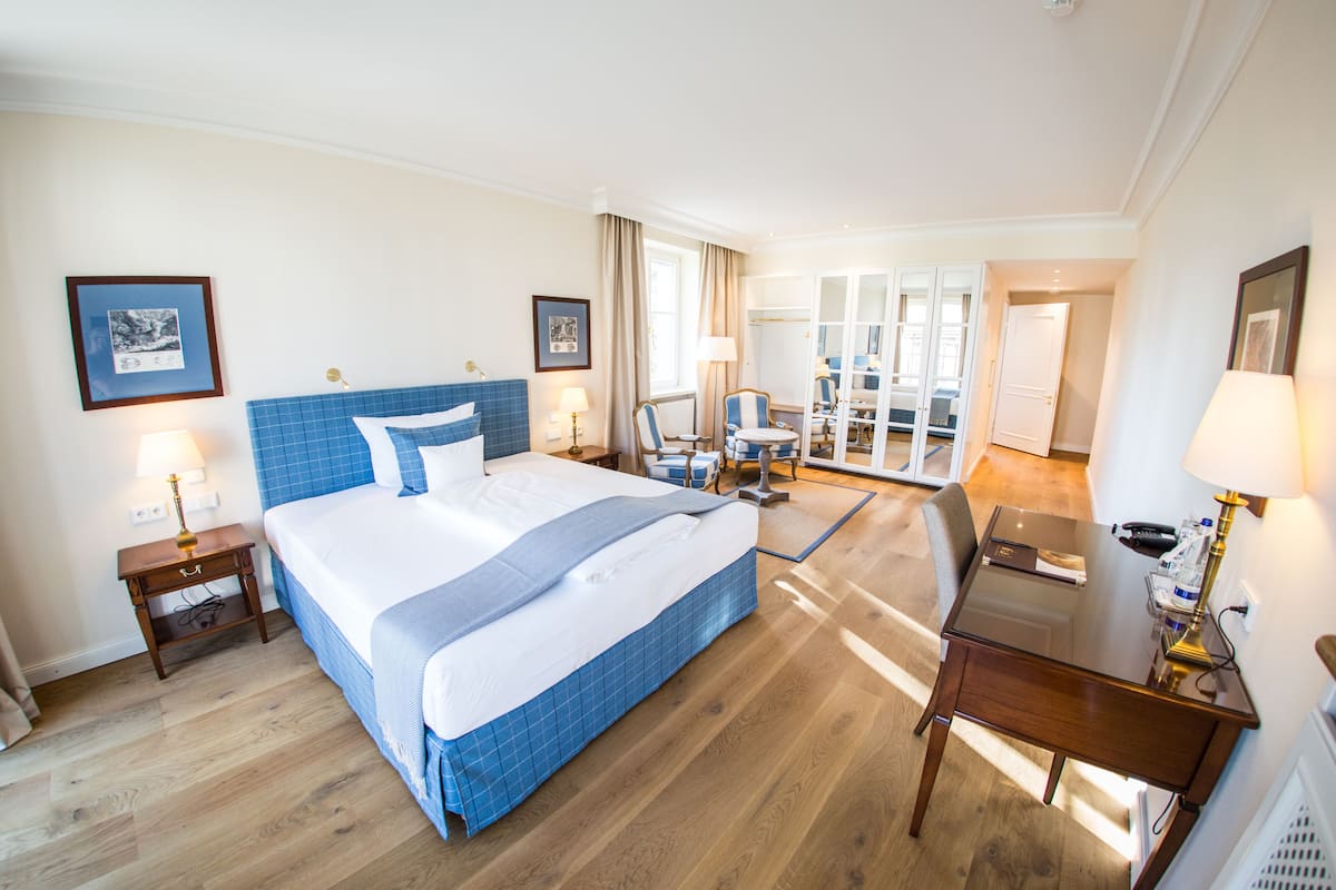 A hotel room with a large bed, a desk, and a chair. There are two lamps on the desk and a chair near the bed.