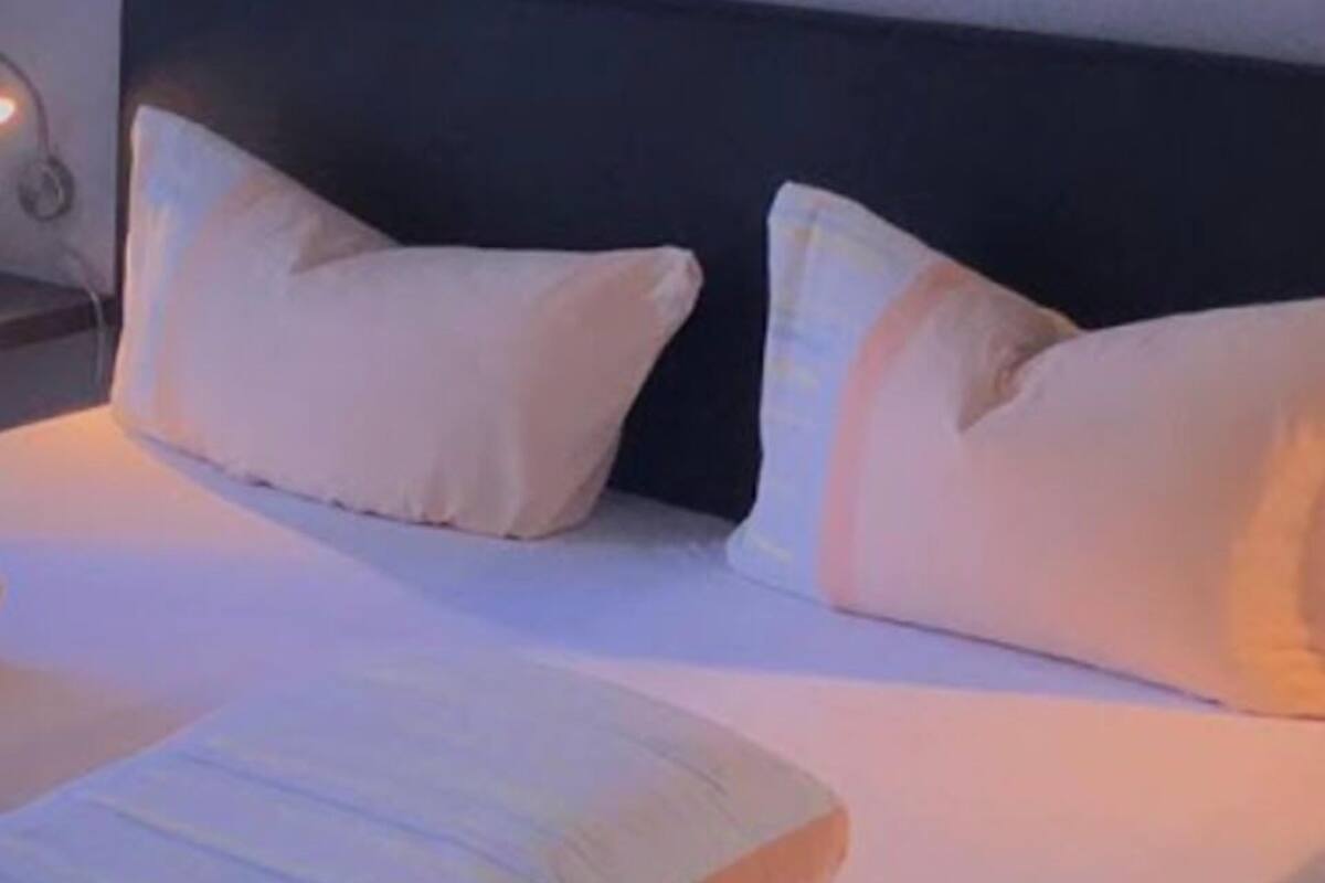 Una cama bien hecha con ropa blanca y dos almohadas, cabecera negra y una lámpara de noche.