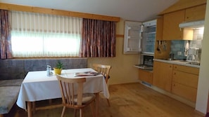 A dining area with a table, chairs, and a bench. There is a window with curtains, a kitchen with cabinets and appliances, and a potted plant on the table.