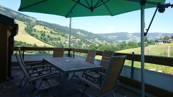 Une terrasse extérieure avec une table et des chaises, un parasol vert et une vue panoramique sur des collines verdoyantes et des maisons.