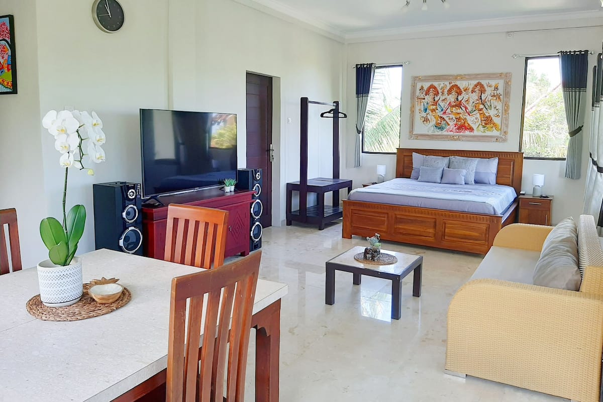 A well-lit room with a bed, sofa, TV, dining table, chairs, and a decorative painting.