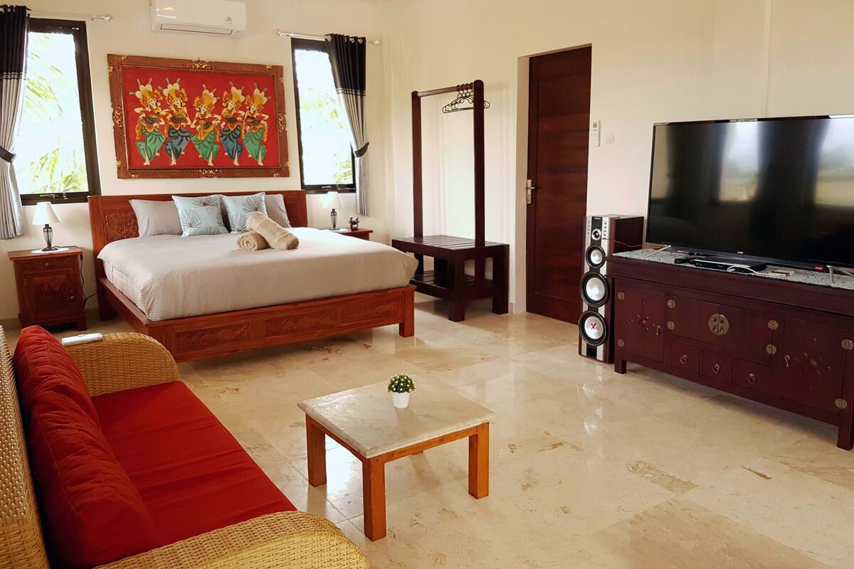 A hotel room with a large bed, a television, a wooden dresser, a small table, and a red sofa.