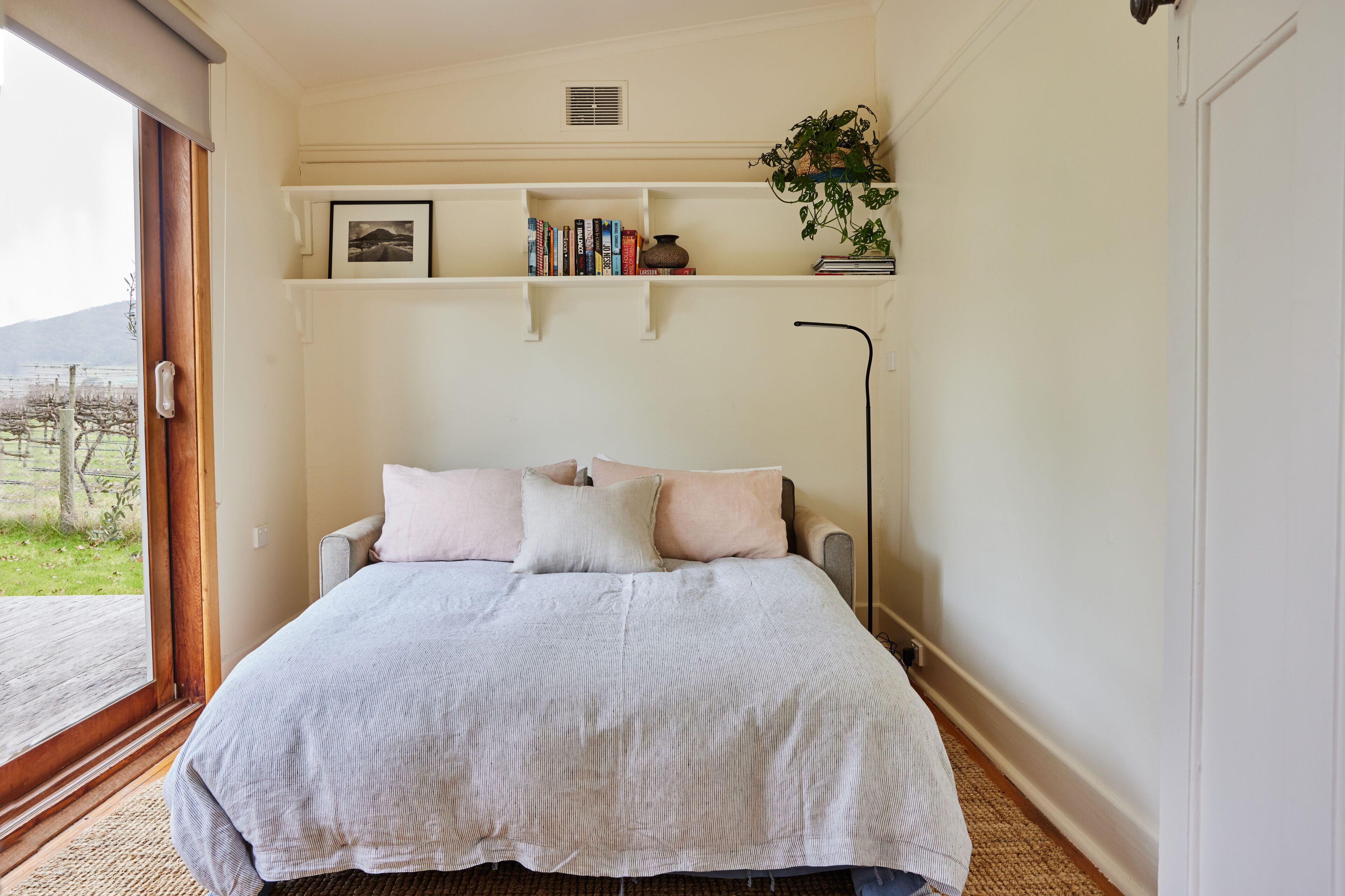 Un dormitorio con cama, una mesita de noche, una estantería con libros y una planta, y una ventana con vistas a la vegetación.