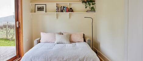 Un dormitorio con cama, una mesita de noche, una estantería con libros y una planta, y una ventana con vistas a la vegetación.