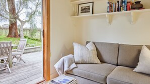 A living room with a sofa, a coffee table with magazines, a bookshelf with books, and a window with a view of a tree and grass. - Yarra Valley Vineyard Cottage - premier location with spectacular views. (Gruyere)