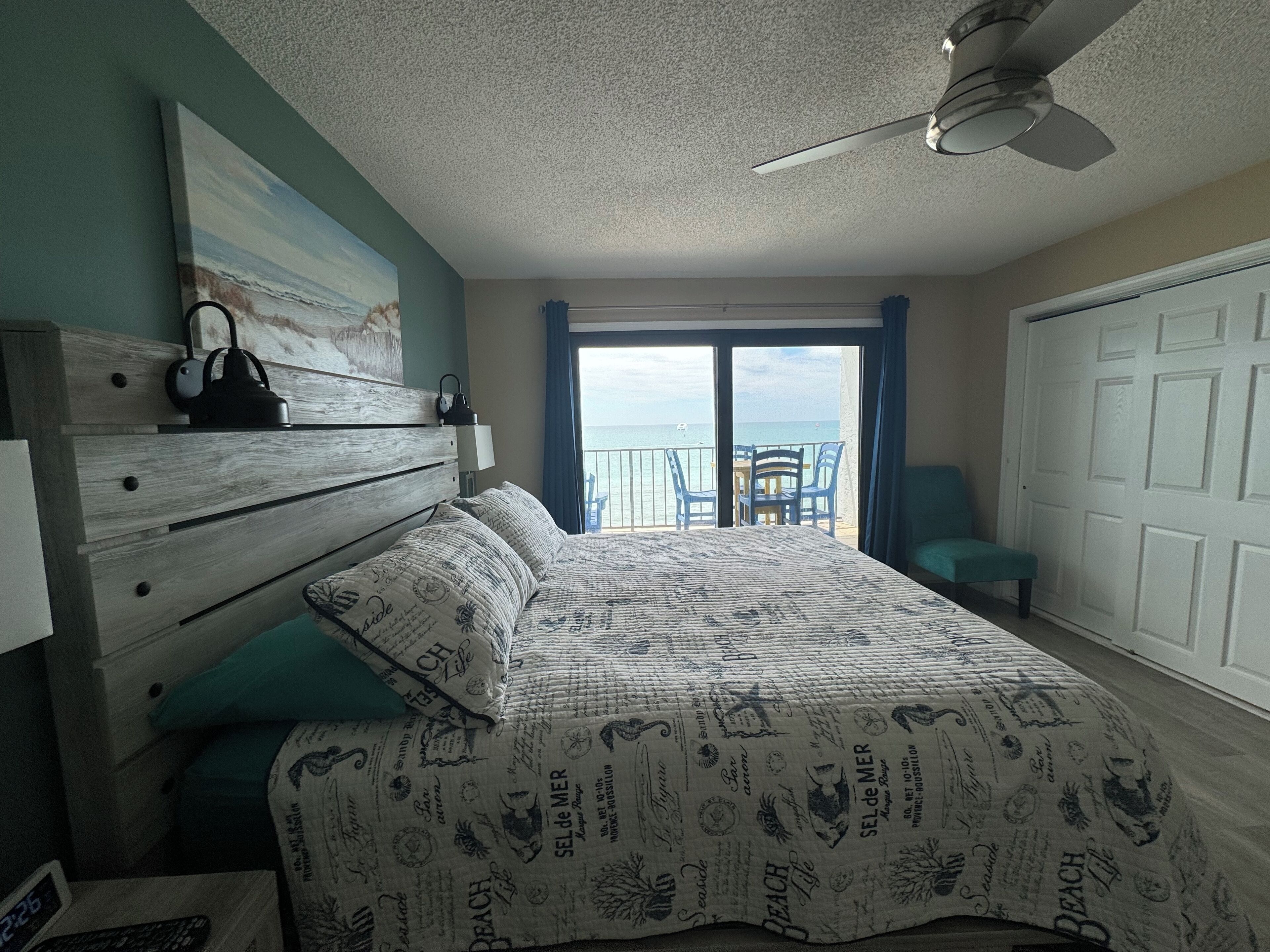 A bedroom with a bed, a nightstand, a ceiling fan, a view of the sea through a sliding door, and a chair by the door.