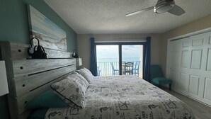 Un dormitorio con cama, mesita de noche, ventilador de techo, vista al mar a través de una puerta corrediza y una silla cerca de la puerta.