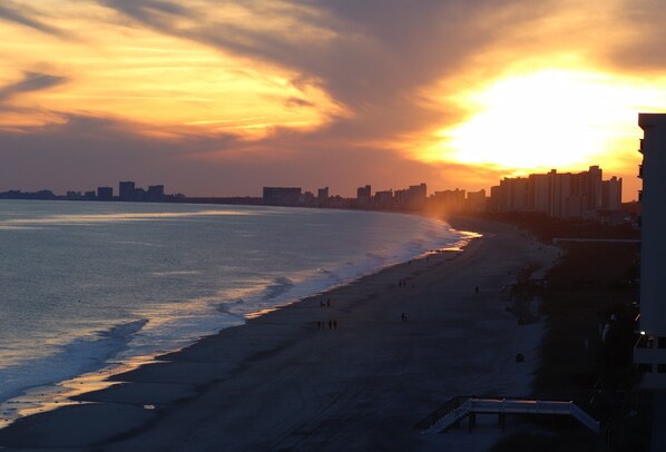 A beach with a sunset, a city skyline in the background, and a few people walking along the shore. - Beautiful Oceanfront 2 Bedroom BOTH OCEANFRONT 2 Bath Condo With AMAZING Views  (North Myrtle Beach)